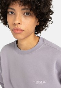 Sudadera lila hecha de un tejido suave, con un cuello redondo acanalado y un logotipo blanco bordado con "ELEMENT CO. SINCE 1992" en el pecho.