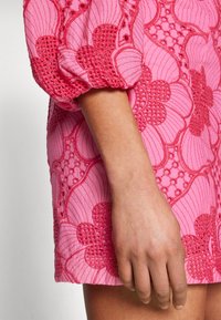 Vestido rosa de mangas curtas com bordados florais, detalhes texturizados e silhueta ajustada, com uma mão visível repousando sobre o tecido.