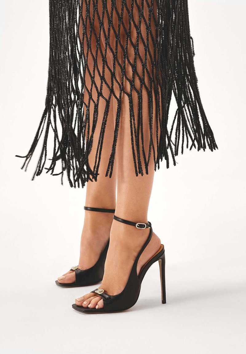 Zwarte open teen hoge hakken hebben een slingback bandje en een gouden accent, met een gladde leren afwerking en een stiletto hak.