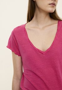 Tricou roz cu decolteu în V, având mâneci scurte cu margini ondulate și un finisaj moale, texturat. Materialul pare ușor și casual.