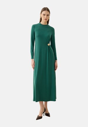 Frau trägt ein langes grünes Kleid mit langen Ärmeln und einem goldenen Verschlussdetail an der Taille, kombiniert mit schwarzen High Heels.