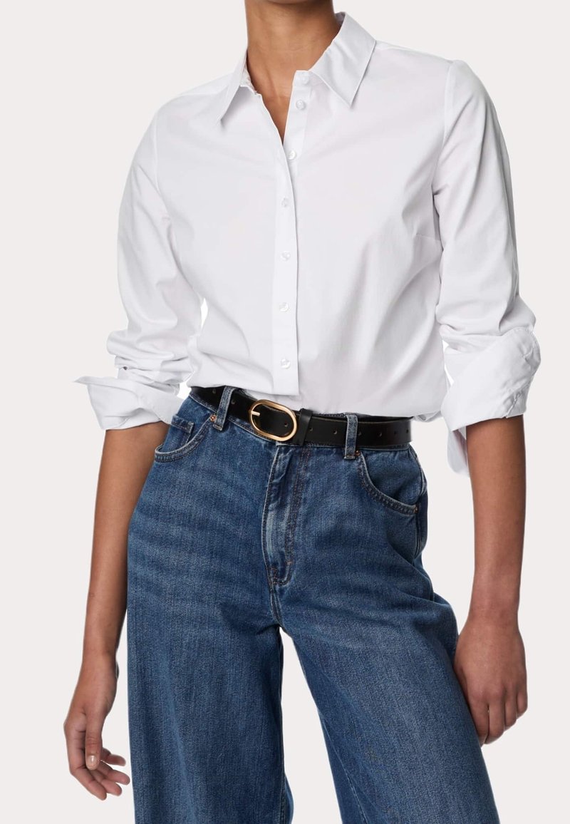 Personne portant une chemise blanche à manches longues boutonnée avec des poignets retroussés, un jean bleu taille haute et une ceinture noire avec une boucle ovale dorée.