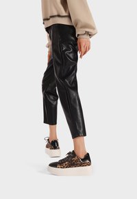 Persona che indossa pantaloni in pelle neri a tre quarti e sneakers con zeppa a stampa leopardata, mostrata dalla parte del polpaccio in giù su sfondo bianco.