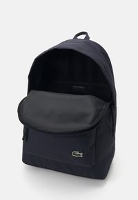 Mochila de tela negra con una parte superior redondeada, gran bolsillo frontal, cierre de cremallera y un pequeño logo de cocodrilo verde en la parte delantera. Interior texturizado.