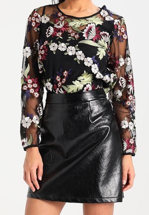 Vrouw draagt een zwarte doorschijnende blouse met bloemenborduursel, gecombineerd met een glanzende zwarte leren minirok.