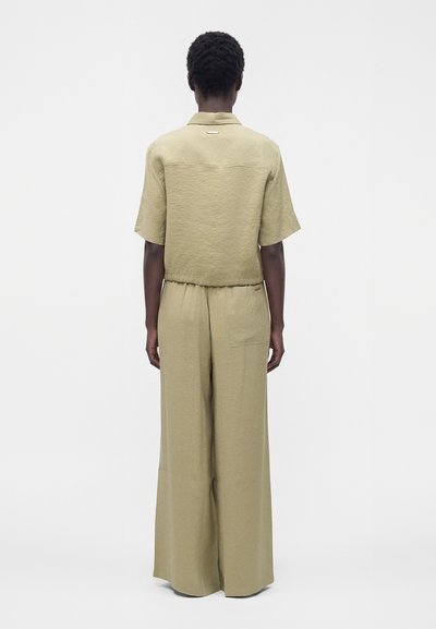 Personne portant une chemise beige à manches courtes et un pantalon à jambes larges, debout de dos sur un fond blanc uni.