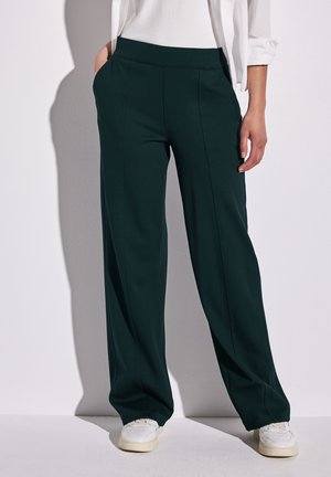 Kobieta ubrana w czarne spodnie z szerokimi nogawkami, białe trampki i białą koszulę z długim rękawem, z podwiniętymi rękawami, stoi na tle jednolitej ściany.