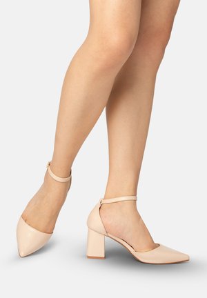 Legs wearing nude pointed-toe block heels with ankle straps on a plain light background.