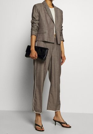 Mujer con blazer de rayas finas color topo y pantalones a juego, llevando un bolso de mano negro, con un top corto blanco y sandalias negras con tacón.