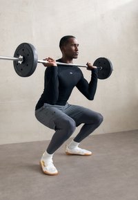 Άνδρας με μαύρη μπλούζα και γκρι κολάν εκτελεί καθίσματα κρατώντας μία μπάρα φορτωμένη στους ώμους του σε έναν μινιμαλιστικό εσωτερικό χώρο.