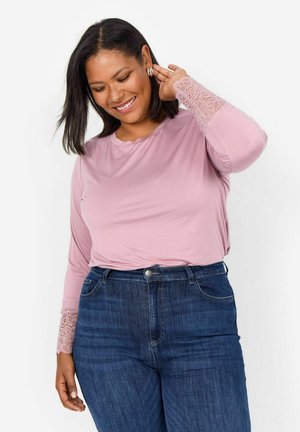 Long-sleeve pink top featuring lace accents on the cuffs, paired with dark blue denim jeans. Smooth fabric with a relaxed fit.