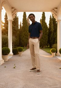 Polo azul-marinho, calças bege e ténis castanhos. Tecido macio, corte casual e marca discreta na camisa. Ambiente exterior com vegetação.