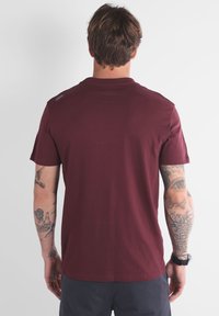 Homme avec des bras tatoués portant un t-shirt bordeaux uni et un short sombre, se tenant de dos face à l'appareil photo sur un fond uni.