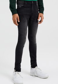 Person som bär svarta skinny jeans, grön långärmad tröja och vita sneakers står framför en enkel ljus bakgrund.
