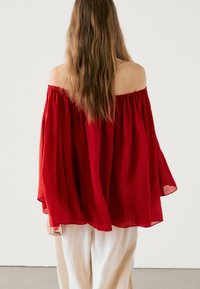 Rød off-the-shoulder bluse med flydende stof, løs pasform og lange ærmer. Letvægts og struktureret materiale, båret med lyse bukser.