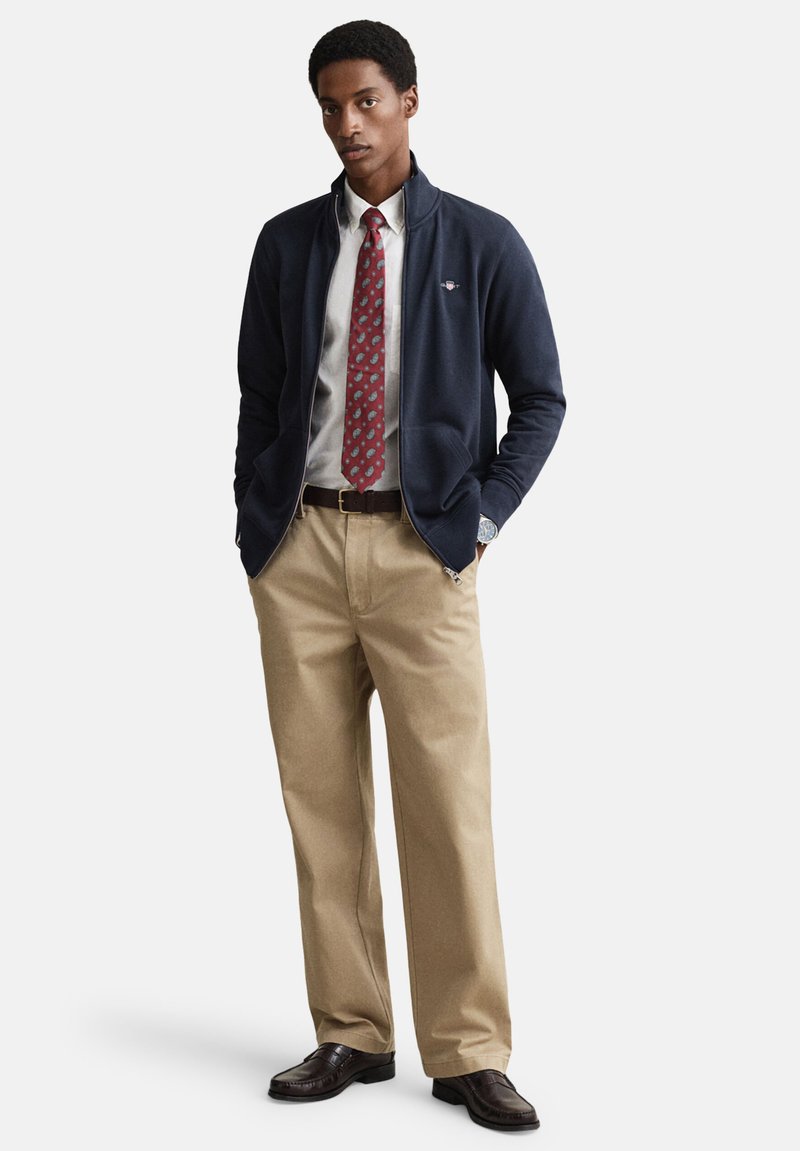 Veste bleu marine zippée portée sur une chemise blanche, cravate rouge à motif, pantalon kaki et chaussures foncées. Détails notables : la veste possède des poches zippées.