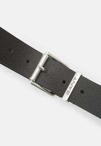 Svart lærbelte med en sølvfarget metallspenne med stangfeste og en polert detalj merket "GANT." Ideelt for ulike antrekk.
