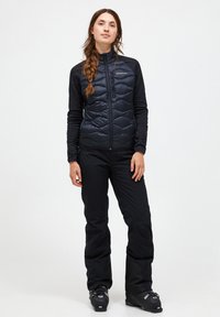 Geïsoleerde zwarte vest met een geborduurd patroon, gedragen met zwarte sneeuwbroek en skischoenen. Aansluitend, lichtgewicht, geschikt voor koude weeractiviteiten.