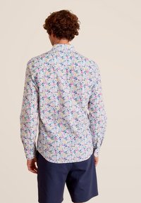 Lange mouw shirt met een witte achtergrond en een bloemmotief in blauw, roze en geel. Met knoopsluiting bij de kraag en een gebogen zoom.