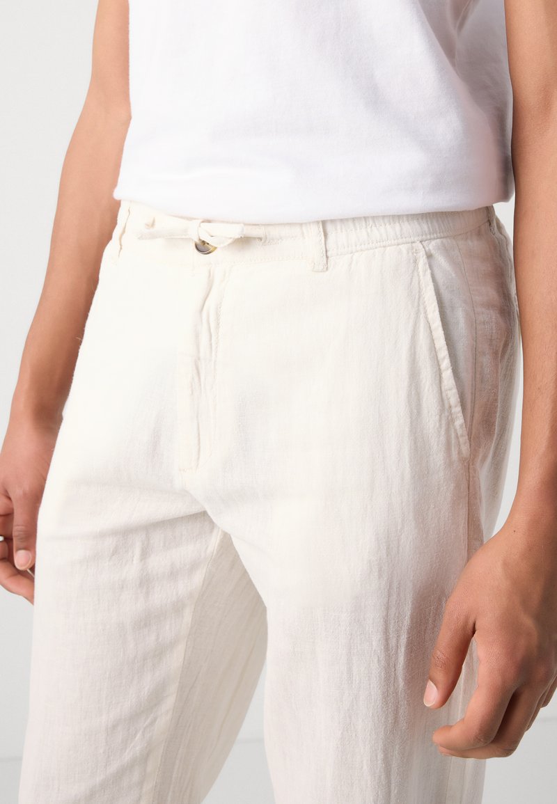 Pantalones de lino crema claro con un ajuste relajado, que presentan una cinturilla elástica, pliegues frontales y bolsillos laterales para mayor comodidad.