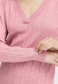 Személy, aki igazít a hosszú ujjú, rózsaszín, bordás kötésű pulóveren, amely V-nyakú.