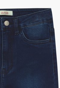 Sección de jeans ajustados de tiro alto en azul oscuro que muestra el bolsillo frontal, el botón, el lazo del cinturón y la etiqueta que dice "720 Tiro Alto Súper Ajustado."