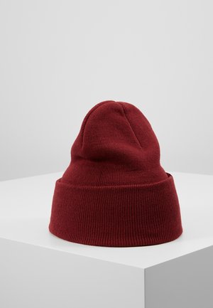 Dunkelroter gestrickter Beanie mit umgeschlagenem Bündchen auf einem weißen Sockel vor hellem grauem Hintergrund.
