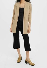Blazer beige chiaro con rever a pupazzo e maniche a tre quarti, abbinato a una maglia nera e pantaloni neri a culotte, completato da scarpe con tacco a blocco color sabbia.