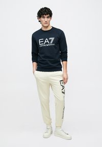 Marineblauwe sweatshirt met wit logo, gecombineerd met crèmekleurige sweatpants met zwarte opdruk. Model draagt witte sneakers.