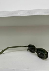 Lunettes de soleil Komono vert foncé avec des verres rectangulaires arrondis reposant sur une surface blanche sous une étagère.