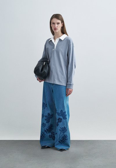 Γκρι μπλούζα πόλο με μακριά μανίκια και λευκό γιακά, συνδυασμένη με φαρδιά μπλε τζιν με λουλουδένια σχέδια. Μαύρη τσάντα ολοκληρώνει την εμφάνιση.