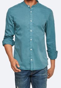 Chemise boutonnée bleu sarcelle avec un col rond, des manches longues retroussées aux poignets et une fermeture à boutons sur le devant, fabriquée en tissu doux et léger.