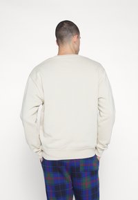 Felpa beige con scollo rotondo, polsini e orlo a costine; presenta una texture liscia. Abbinata a pantaloni a quadri in blu scuro, verde e rosso.