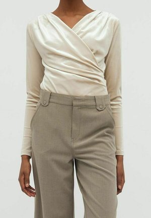 Personne portant un haut cache-cœur à manches longues de couleur crème, rentré dans un pantalon beige taille haute avec des détails de patte à boutons à la ceinture.