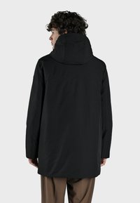 Chaqueta con capucha negra de textura suave, mangas largas y corte recto, con un diseño minimalista y sin detalles de cierre visibles.