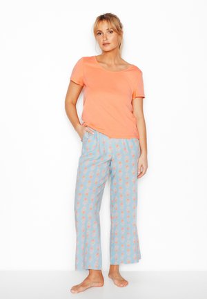 Donna in piedi scalza che indossa una maglietta arancione a maniche corte e pantaloni larghi azzurri con un motivo floreale arancione.