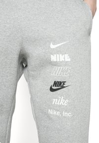 Grå träningsoveraller med olika Nike-logotyper i svart och vitt. Materialet är en mjuk bomullsblandning; designen inkluderar screentryckt text och symboler.