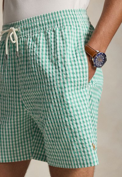 Rohelised ja valged ruudulised lühikesed püksid koos paelaga, inimese käsi taskus, randmel sinise sihverplaadiga kell pruunist nahast rihmaga.