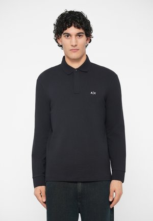 Jovem com cabelo preto encaracolado a usar uma polo de manga longa preta com um pequeno logótipo "A|X" no lado esquerdo do peito.