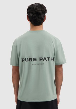 Man met donker krullend haar van achteren gezien, die een lichtgroen T-shirt draagt met "PURE PATH ATHLETICS CLUB" in het zwart gedrukt op de achterkant.