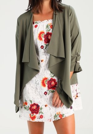 Vrouw draagt een witte kanten jurk met rood- en oranje bloemenpatronen en een open olijfgroene gewadenjas met knoopmouwen.