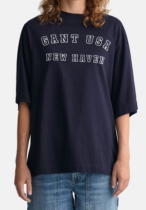Person, der ein großes navy T-Shirt mit dem Aufdruck "GANT USA NEW HAVEN" trägt, kombiniert mit hellblauen Jeans.