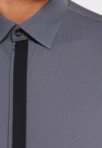 Camicia grigia con colletto abbottonato, caratterizzata da una striscia verticale nera al centro e un piccolo logo grigio in basso a destra.