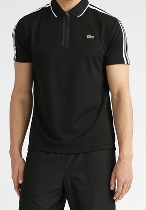 Mann trägt ein schwarzes Polo-Shirt mit kurzen Ärmeln, weißen Streifen an den Ärmeln, einem Reißverschluss am Kragen und einem kleinen grünen Krokodil-Logo auf der Brust.