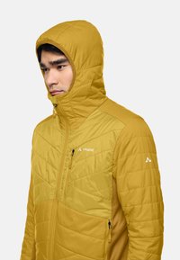 Veste isolante en polyester jaune avec capuche, dotée d'une poche poitrine zippée, de coutures en diagonale et d'une texture lisse.