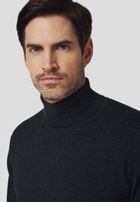 Mężczyzna z krótkimi ciemnymi włosami i jasnym zarostem, ubrany w czarny golf, na jednolitym jasnym tle.