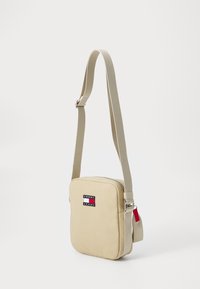 Beige lerret crossover-veske med justerbar stropp, firkantet form, glidelås-lukking og et logo-lapp med røde, hvite og blå detaljer.