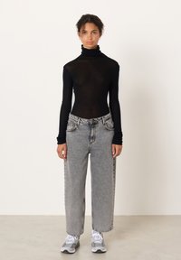 Noor inimene seisab musta läbipaistva kõrge kaelusega särgi, lahtiste hallide teksade ja hallide tossudega ühetaustalise heleda tausta ees.