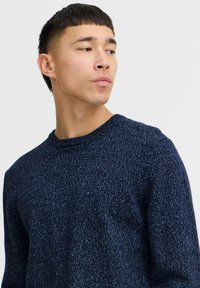 Marineblå strikkegenser med rund hals, med et teksturert mønster og ribbet kant, vist på en mannlig modell mot en ensfarget bakgrunn.