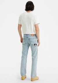 Vaqueros desgastados de color azul claro con un parche de signo de paz en el bolsillo trasero, combinados con una camiseta de manga corta blanca y zapatos beige.